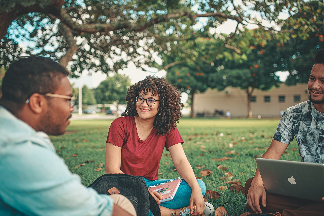 groupe d'étudiants dans un parc avec un cahier et un ordinateur entrain de se regarder et sourire